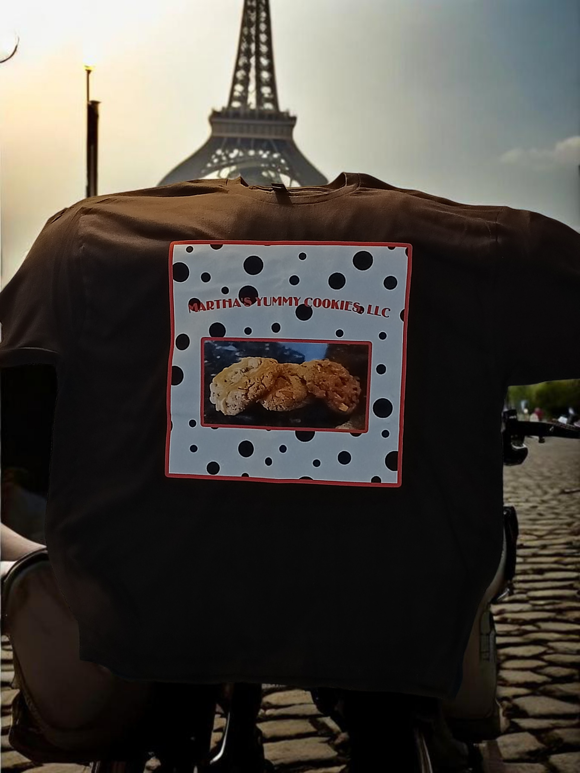 A brown t-shirt featuring a graphic of cookies, worn by someone on a cobblestone path in front of the Eiffel Tower.