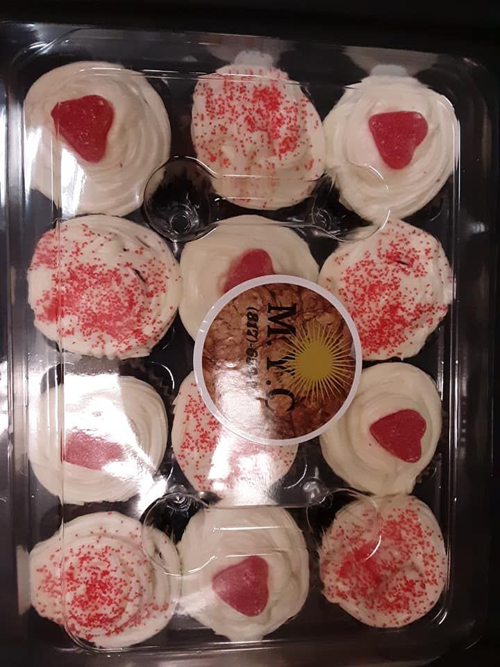A dozen white frosted cupcakes with red sprinkles and strawberry hearts in a clear plastic container.