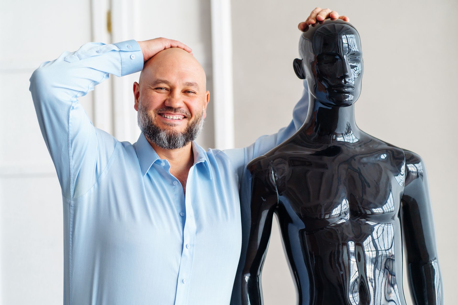 Smiling man in a light blue shirt standing beside a black mannequin, hand on head, in a bright room