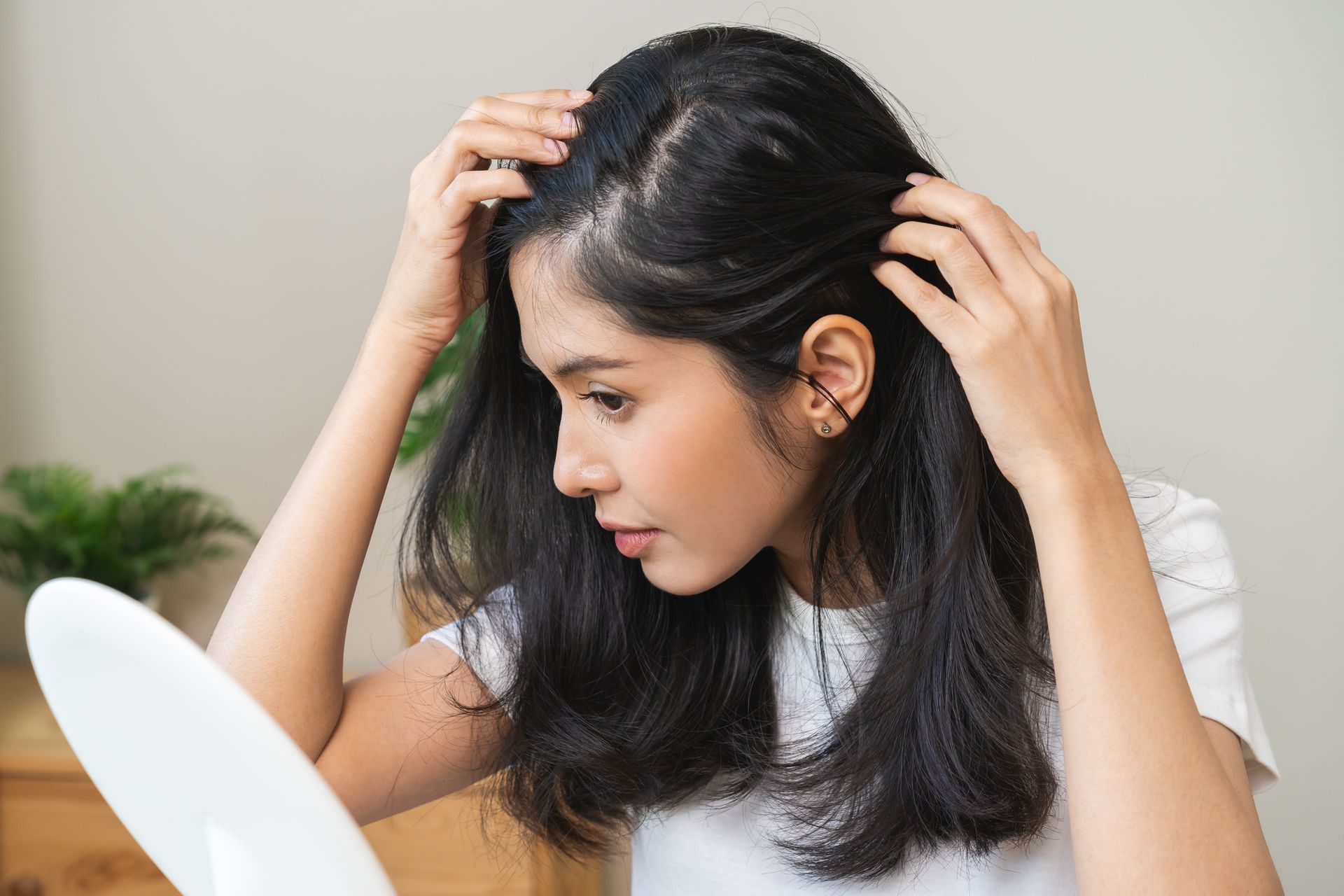 Woman parting her hair with both hands to examine her scalp in a mirror.