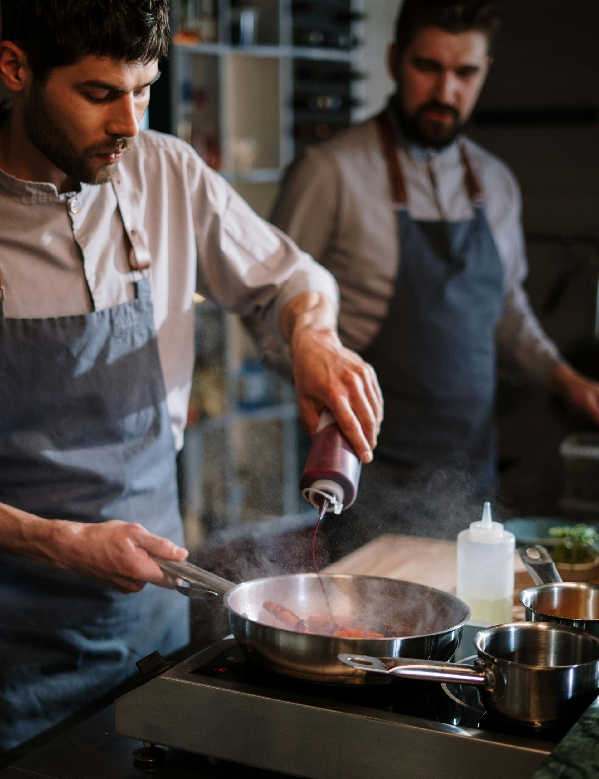 Two men are cooking in a kitchen and one of them is adding pepper to a pan.