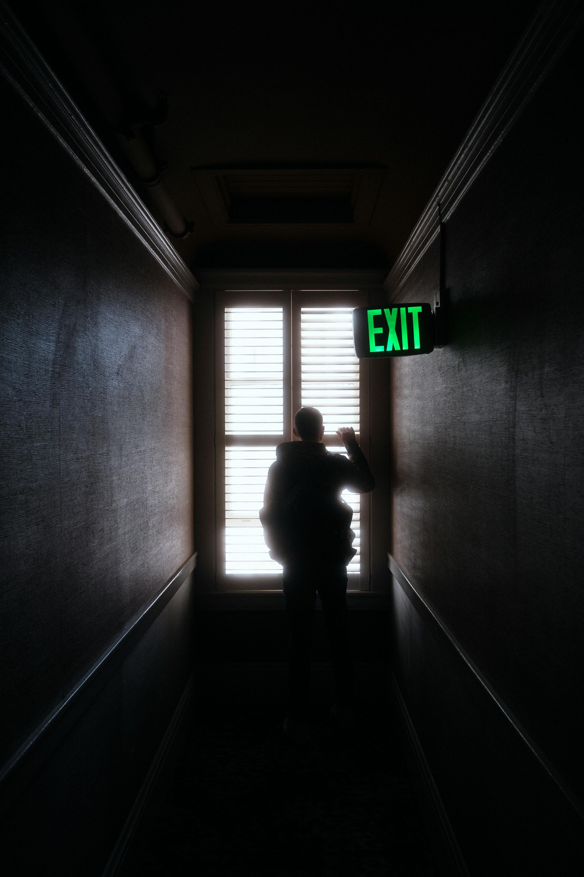 A person is standing in a dark hallway next to an exit sign