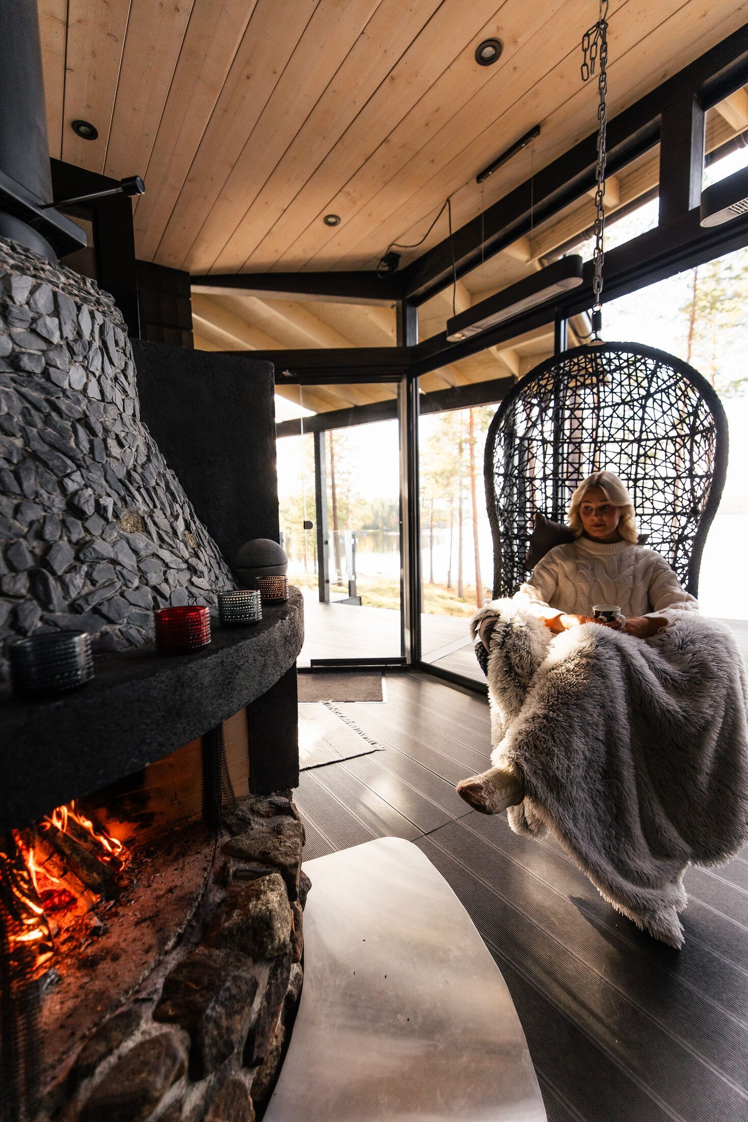 Guest wrapped in a blanket in a hanging chair next to a stone fireplace in a Lapland holiday home
