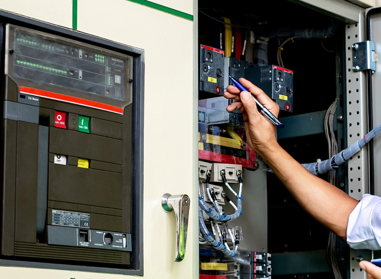 Technician using a multimeter to inspect an open electrical control cabinet beside a panel display