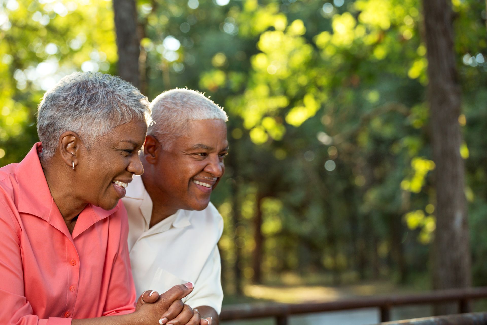 Two smiling people leaning together outdoors in a sunlit park.