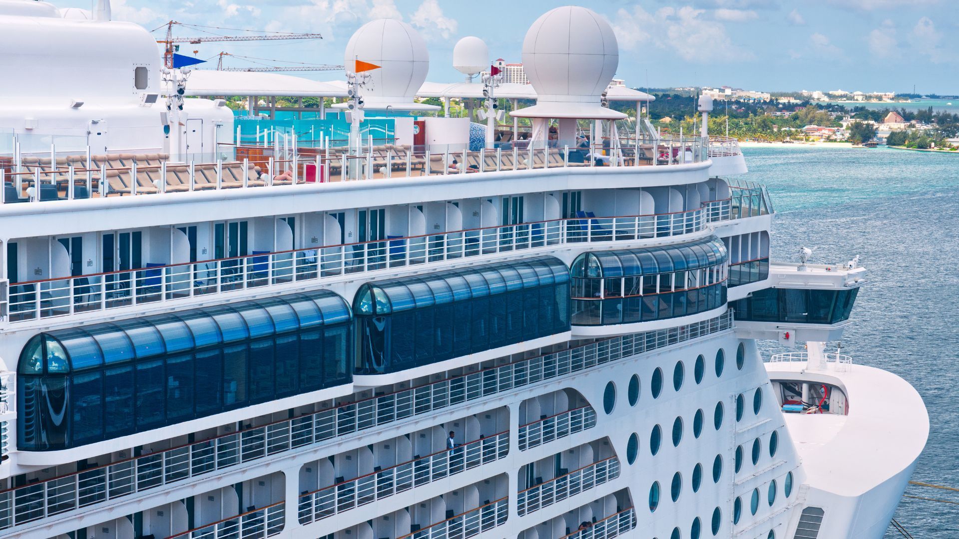 Cruise ship deck with white radar domes and blue-tinted windows, docked near a coastal city