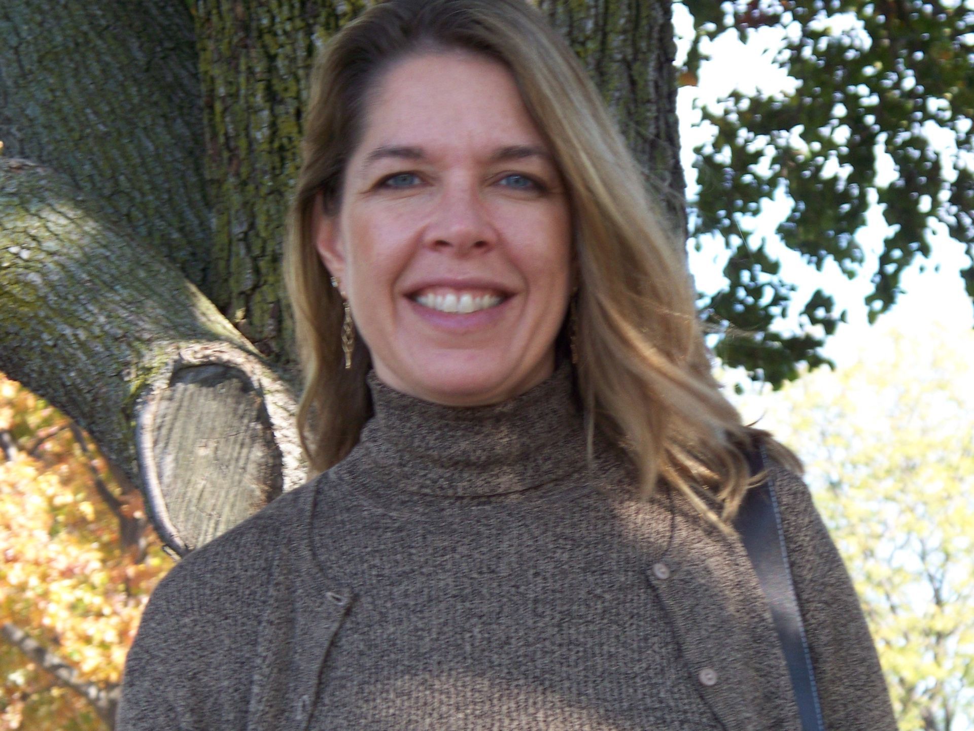 Krystene Rivers in a brown sweater is smiling in front of a tree
