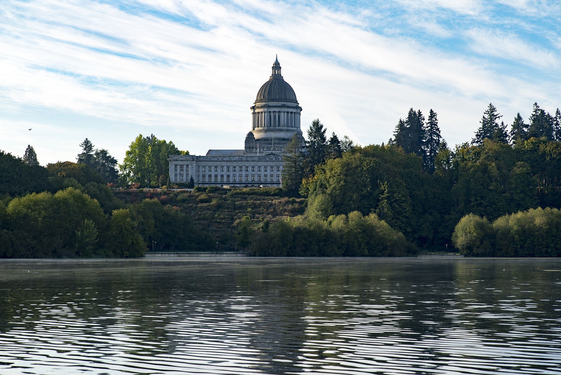 A large building is sitting on top of a hill overlooking a lake surrounded by trees.