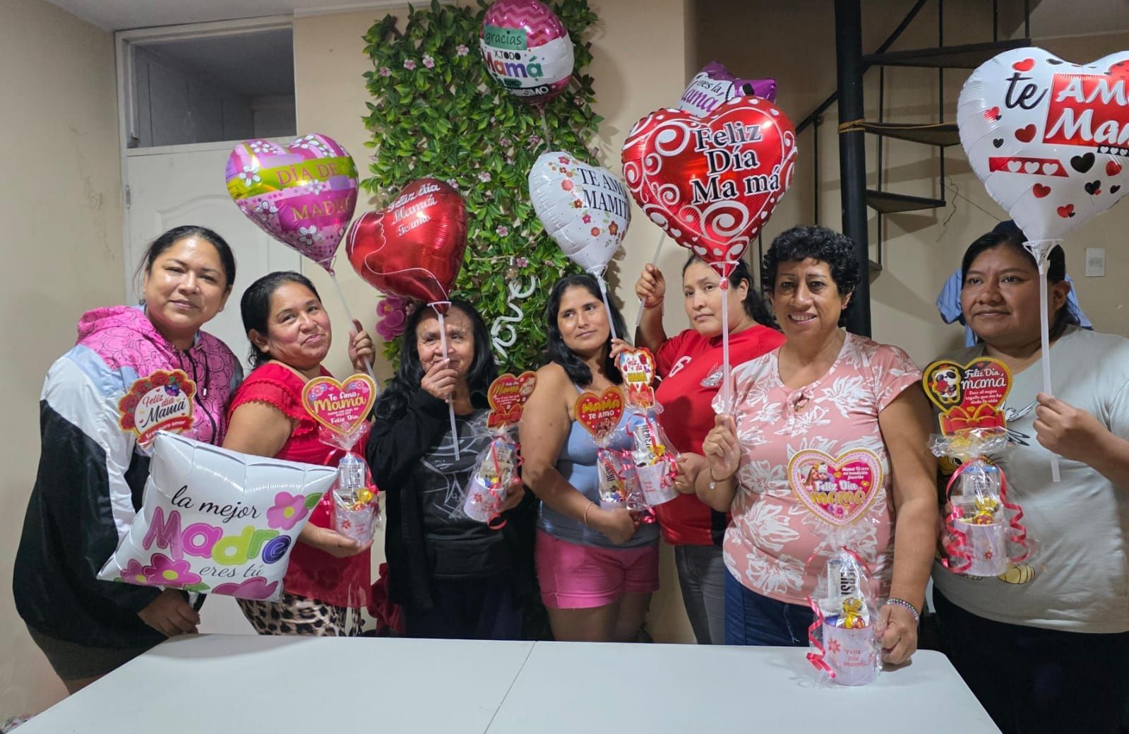 Un grupo de personas, con globos de colores y regalos, sonríen en una celebración en el interior de un edificio.