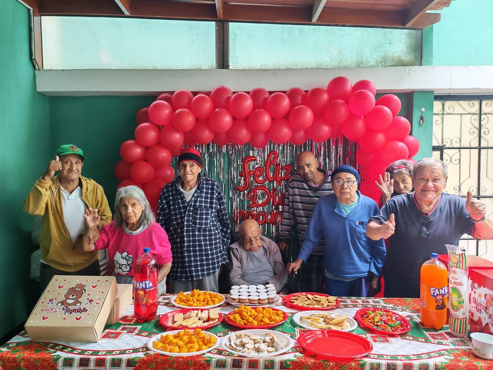 Grupo posando detrás de una mesa de cumpleaños con un arco de globos rojos y pasteles en un espacio exterior cubierto.