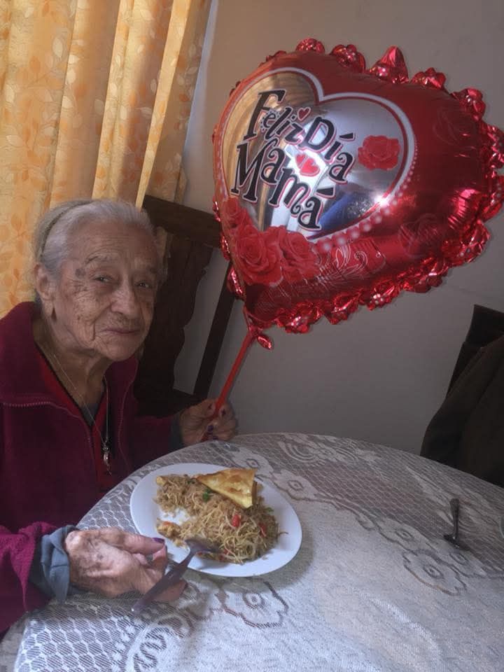 Anciana sentada a una mesa con un pastel, junto a un globo rojo en forma de corazón que dice “Feliz Cumpleaños”.