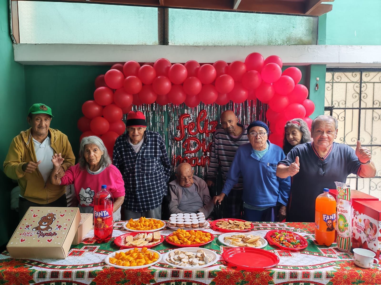 Grupo posando detrás de una mesa con comida y bebidas frente a un fondo de globos rojos en una celebración.
