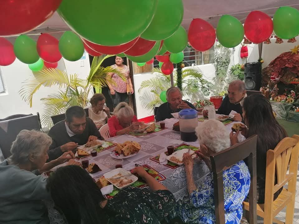 Personas sentadas en una mesa festiva al aire libre, bajo globos rojos y verdes, compartiendo una comida.