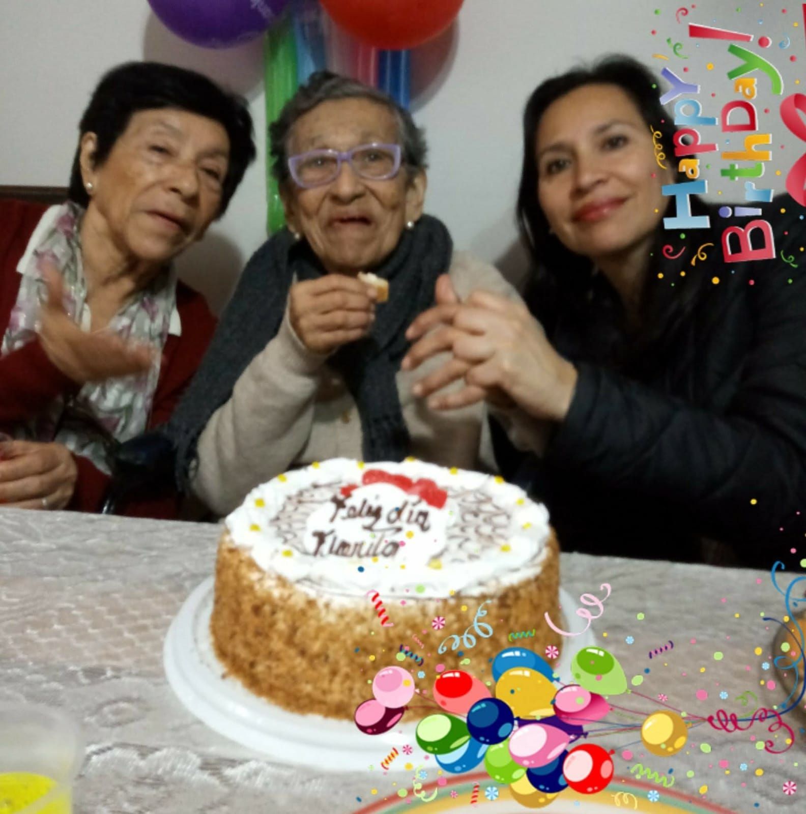 Tres personas sonriendo detrás de una tarta de cumpleaños con globos y coloridas decoraciones de fiesta.