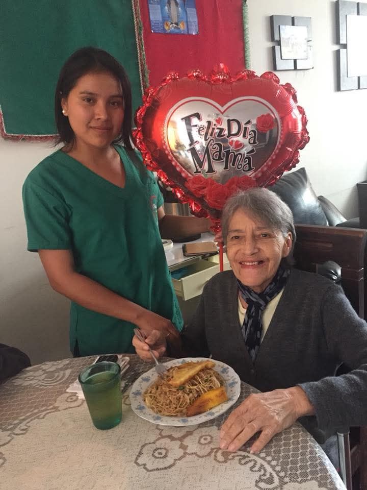 Una mujer está de pie junto a un anciano sentado a la mesa con comida y un globo en forma de corazón que dice 