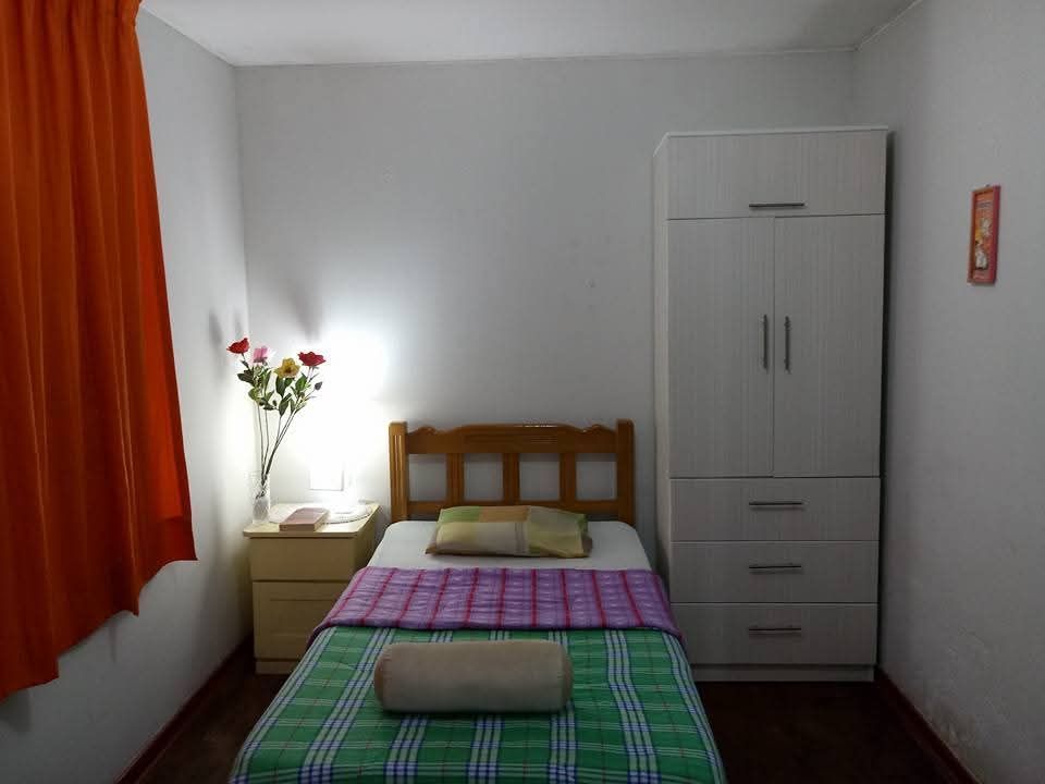 Habitación pequeña con dos camas individuales, armario blanco, cortinas naranjas y un jarrón con flores junto a la ventana.