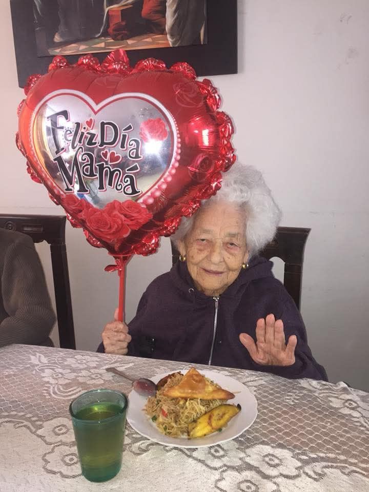 Una anciana sentada a una mesa sostiene un globo de San Valentín con forma de corazón junto a un plato de comida.