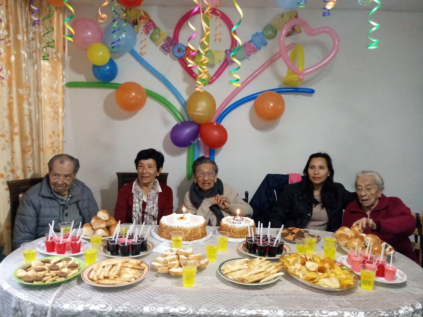 Seis personas sentadas en una mesa de cumpleaños decorada con pasteles, bocadillos y globos de colores detrás.