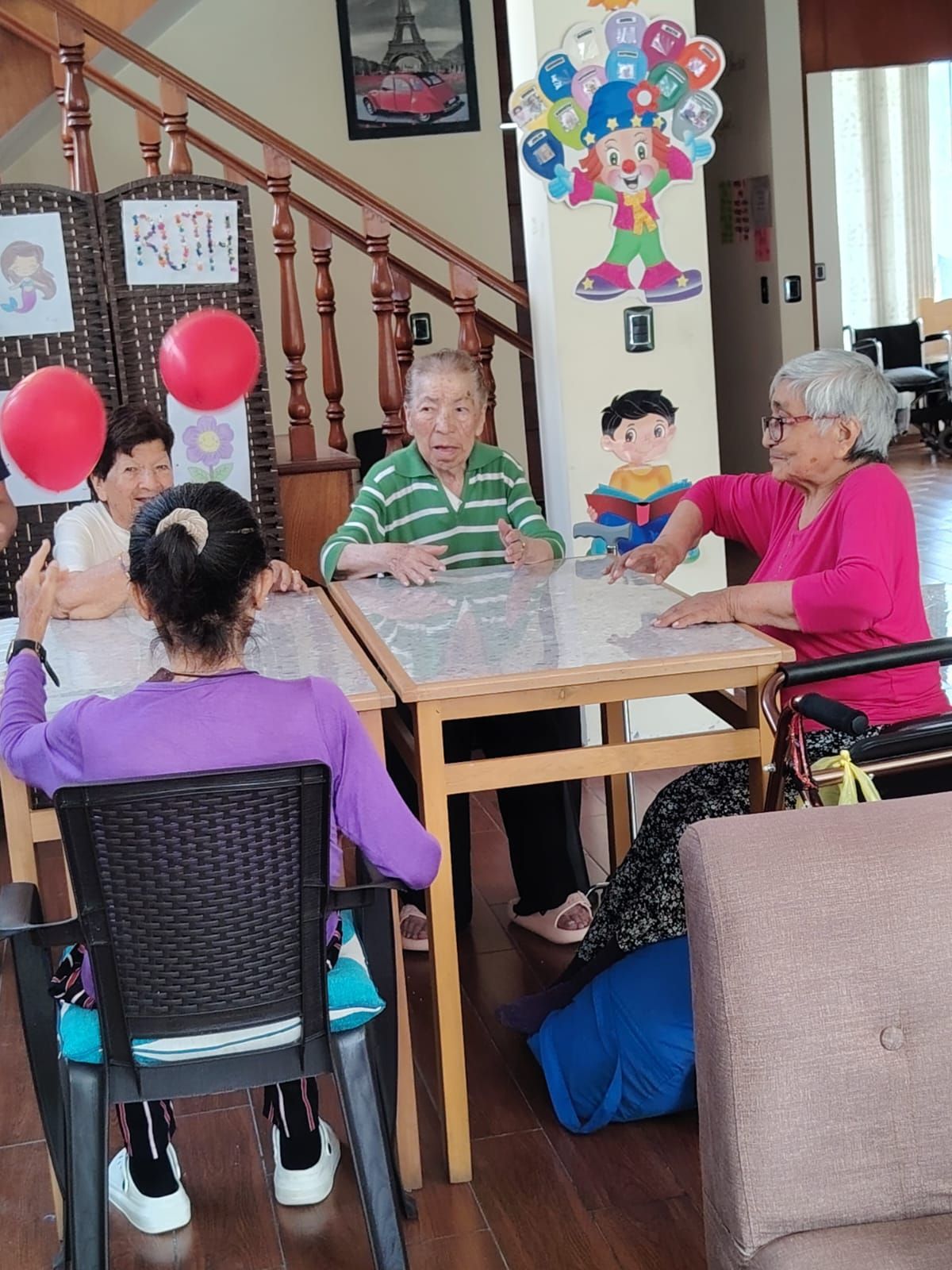 Varias personas sentadas alrededor de una mesa en una casa, con globos y una colorida decoración en la pared cerca.