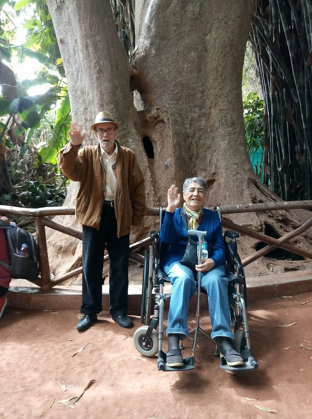 Dos personas frente a un árbol gigante, una de pie saludando con la mano, la otra sentada en una silla de ruedas.