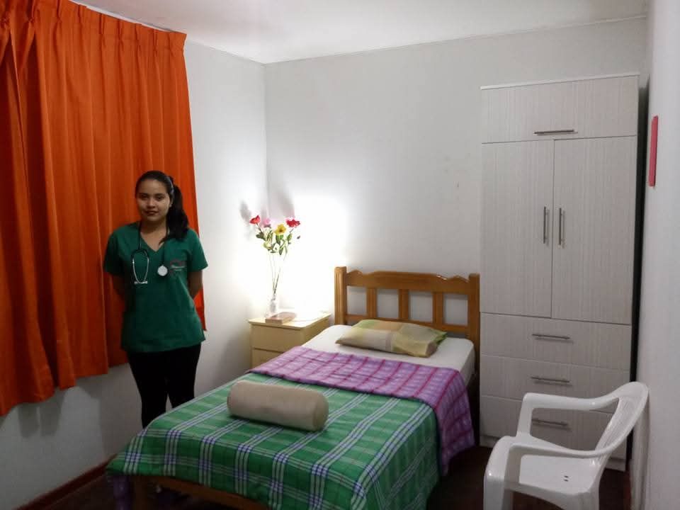 Dormitorio con una mujer de pie junto a una ventana con cortinas naranjas, una cama y un armario blanco.