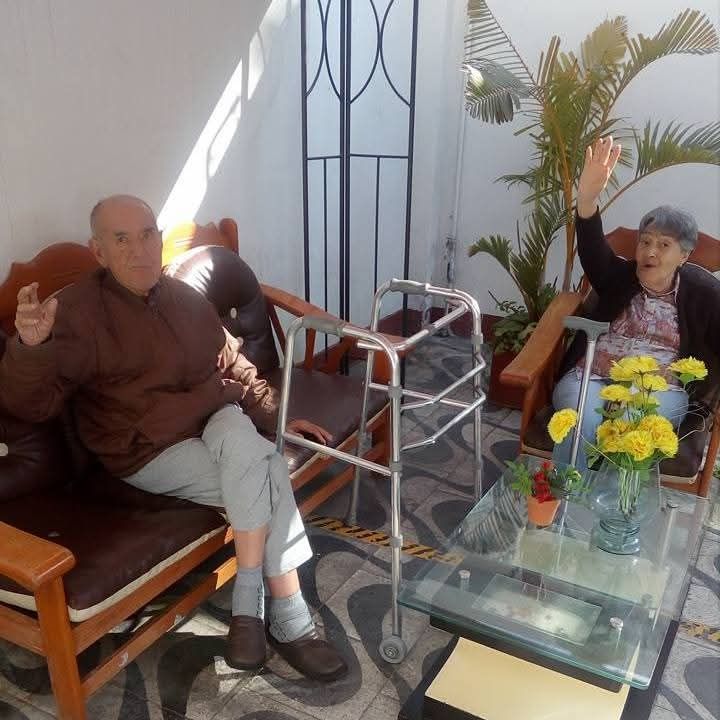 Dos personas están sentadas en un luminoso salón de patio con un andador y una mesa de cristal con flores.