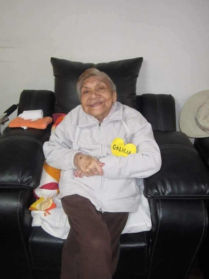 Persona sonriente sentada en un sofá de cuero negro, con una chaqueta clara y una etiqueta amarilla con su nombre.