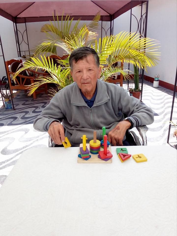 Un hombre está sentado en una mesa con coloridos juguetes apilables bajo un patio sombreado.