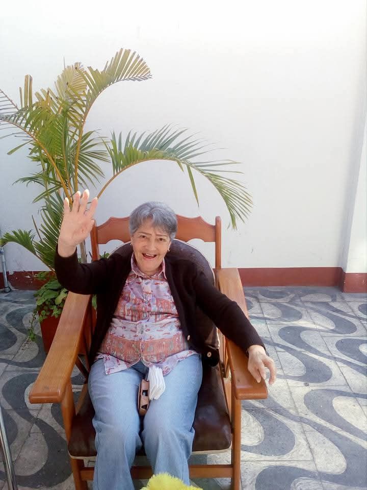 Una persona sonriente sentada en una silla de madera saluda junto a una palmera en maceta en una habitación luminosa.
