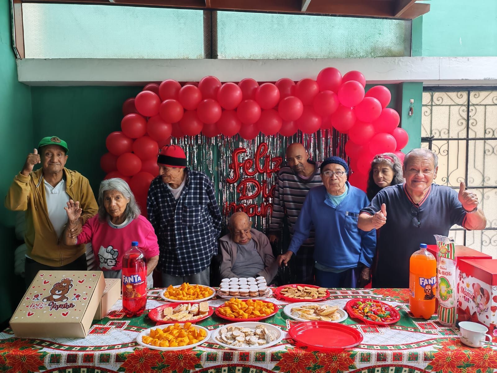 Un grupo posando detrás de una mesa con coloridos platos de comida en una celebración decorada con globos rojos.