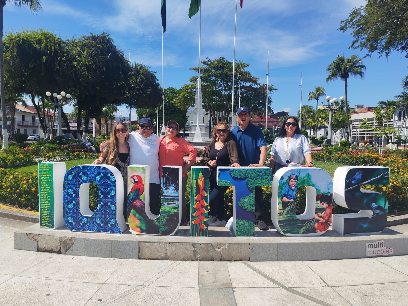 Grupo posando detrás de las coloridas letras “IQUITOS” en una soleada plaza con palmeras y cielo azul.
