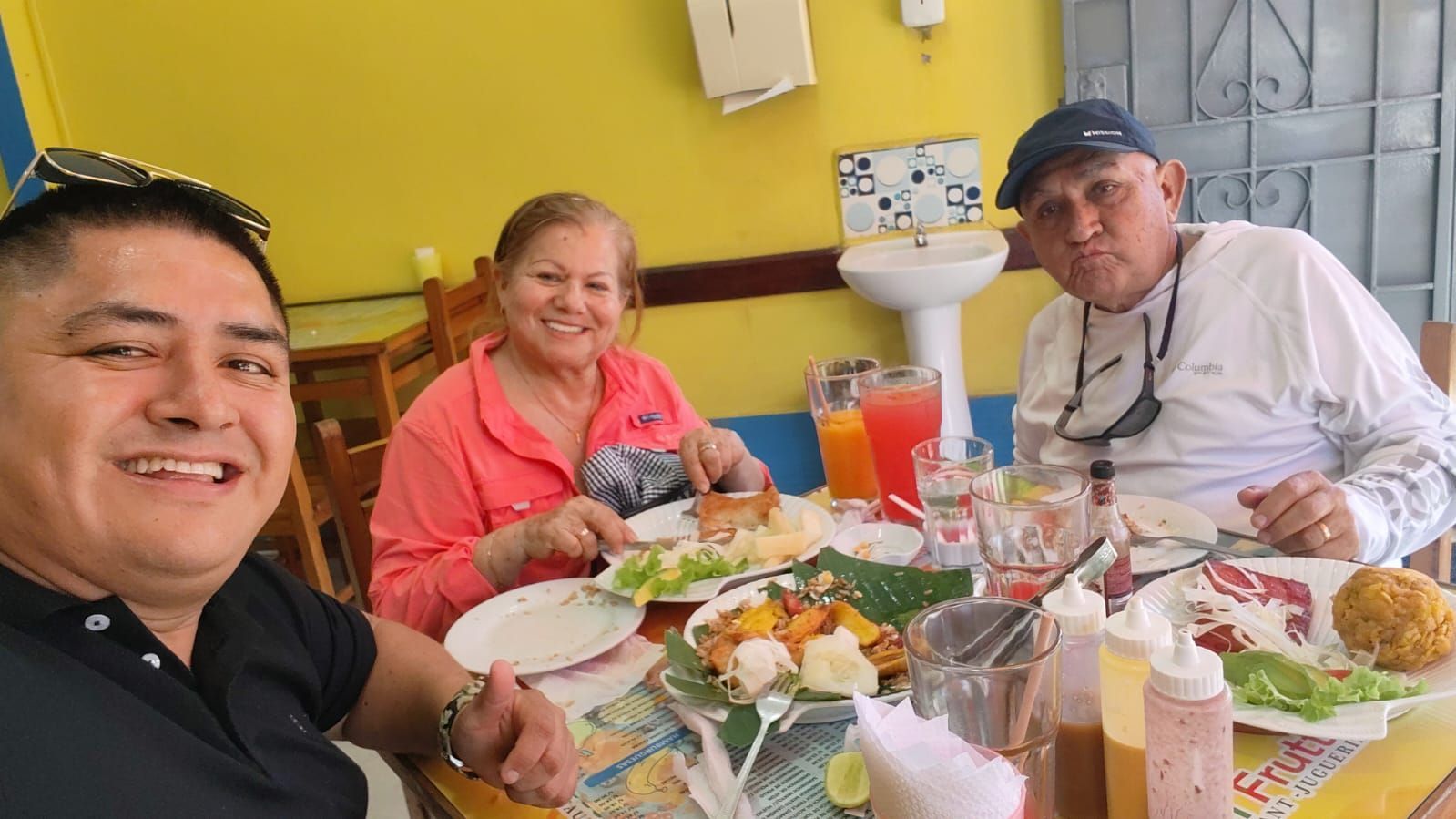 Tres personas sonriendo en una mesa con comida y bebidas en un comedor de color amarillo brillante.