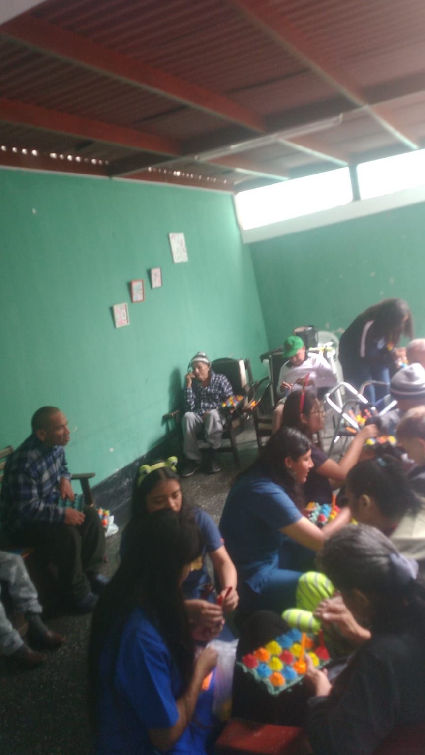 En una sala con paredes verdes, varias personas estaban sentadas, reunidas alrededor de una mesa con objetos coloridos.