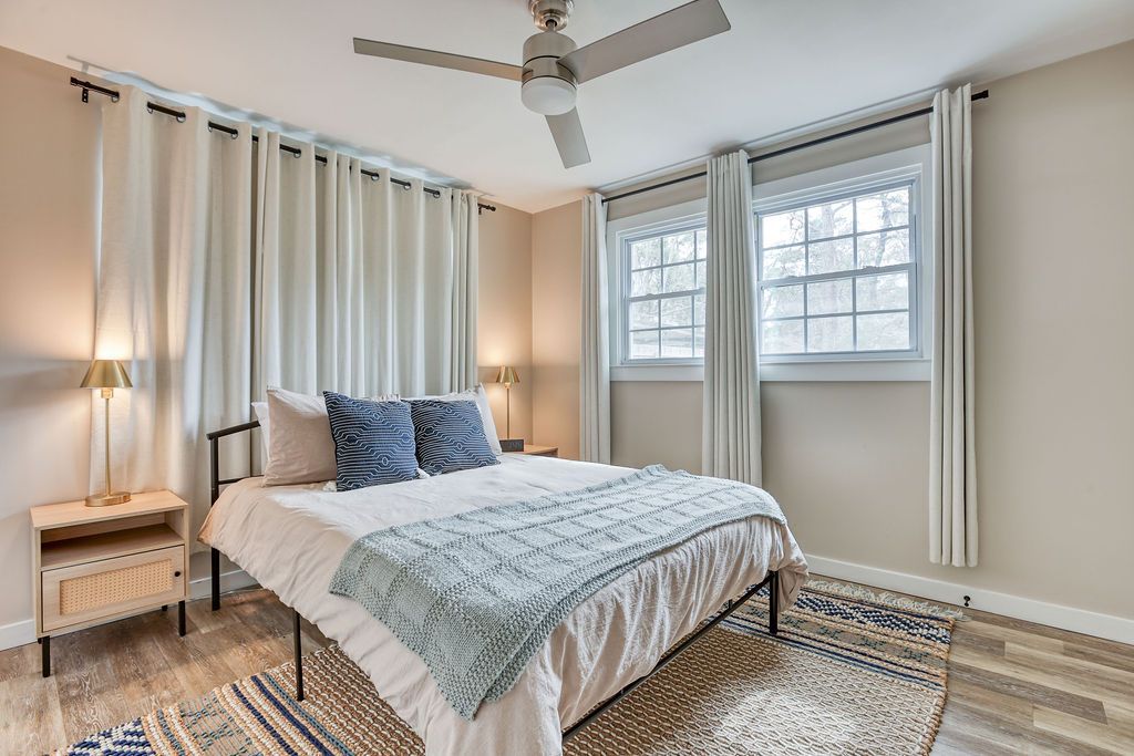 Bedroom with bed, nightstands, windows, and light-colored walls and curtains; a ceiling fan is overhead.
