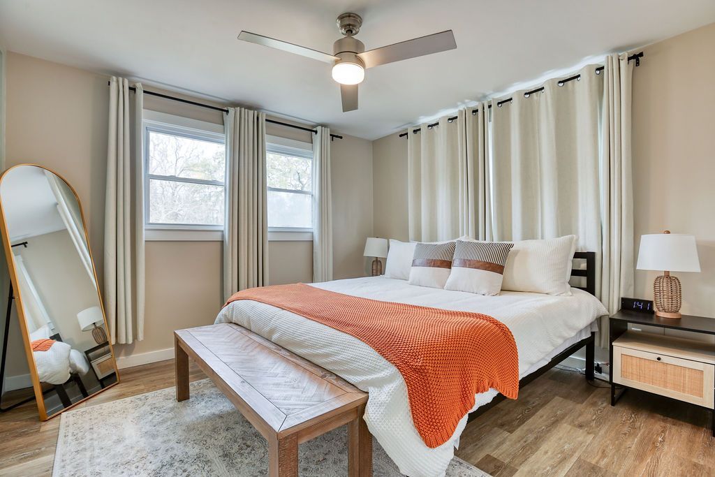 Bedroom with bed, bench, mirror, windows, curtains, and rug; orange and neutral tones.
