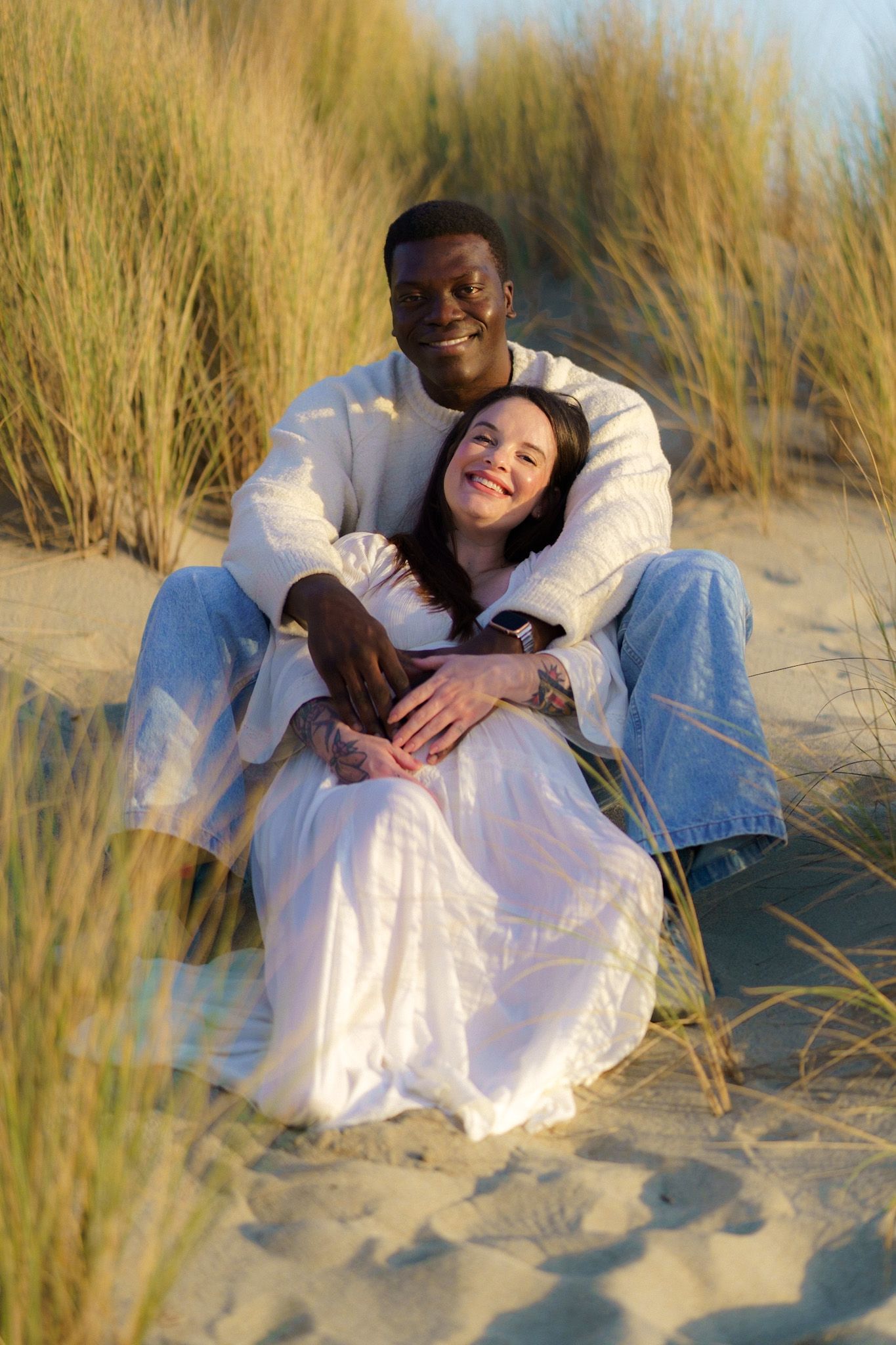 Couple embracing on a sandy dune, surrounded by tall grass. Man in white sweater, woman in white dress.