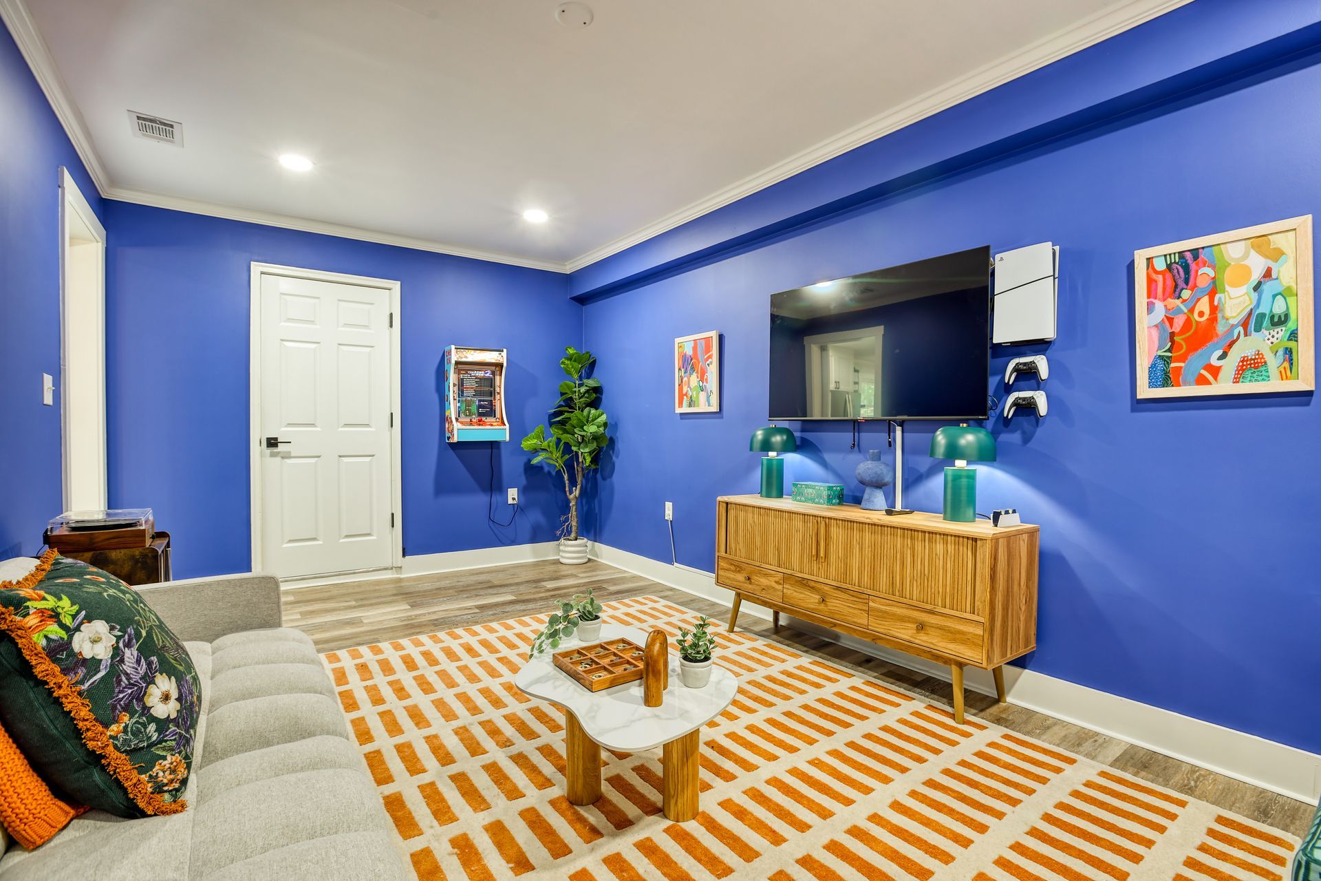 Blue-walled living room with a gray couch, patterned rug, wooden console, and TV. Artwork and a faux plant are present.