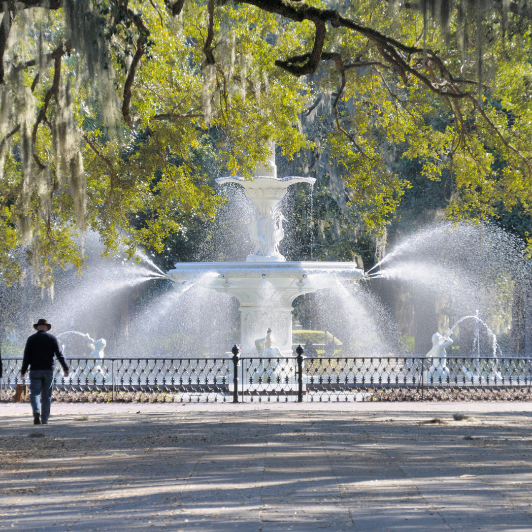 White fountain sprays water in a park, person walks in front of it.