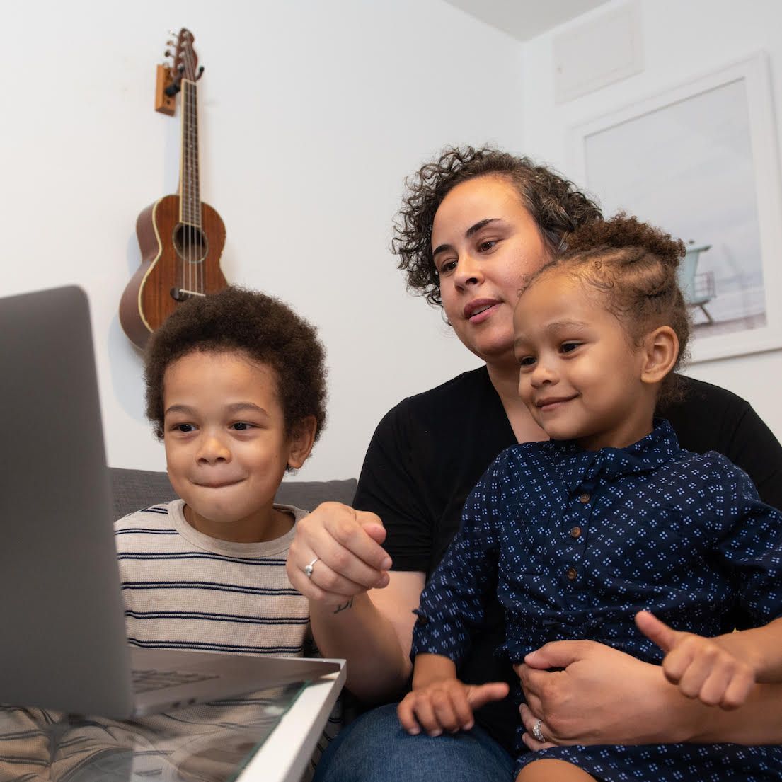Three people gathered around a laptop, smiling in a bright room with a guitar on the wall.