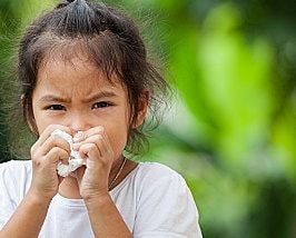 Child blowing nose with a tissue, looking uncomfortable, against a blurred green background