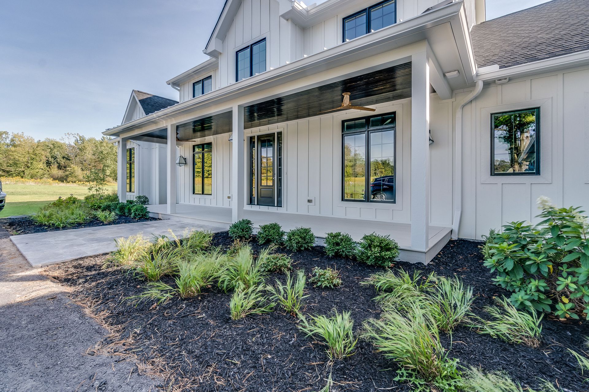 White farmhouse with black-framed windows and front porch, featuring landscaping with dark mulch and greenery.