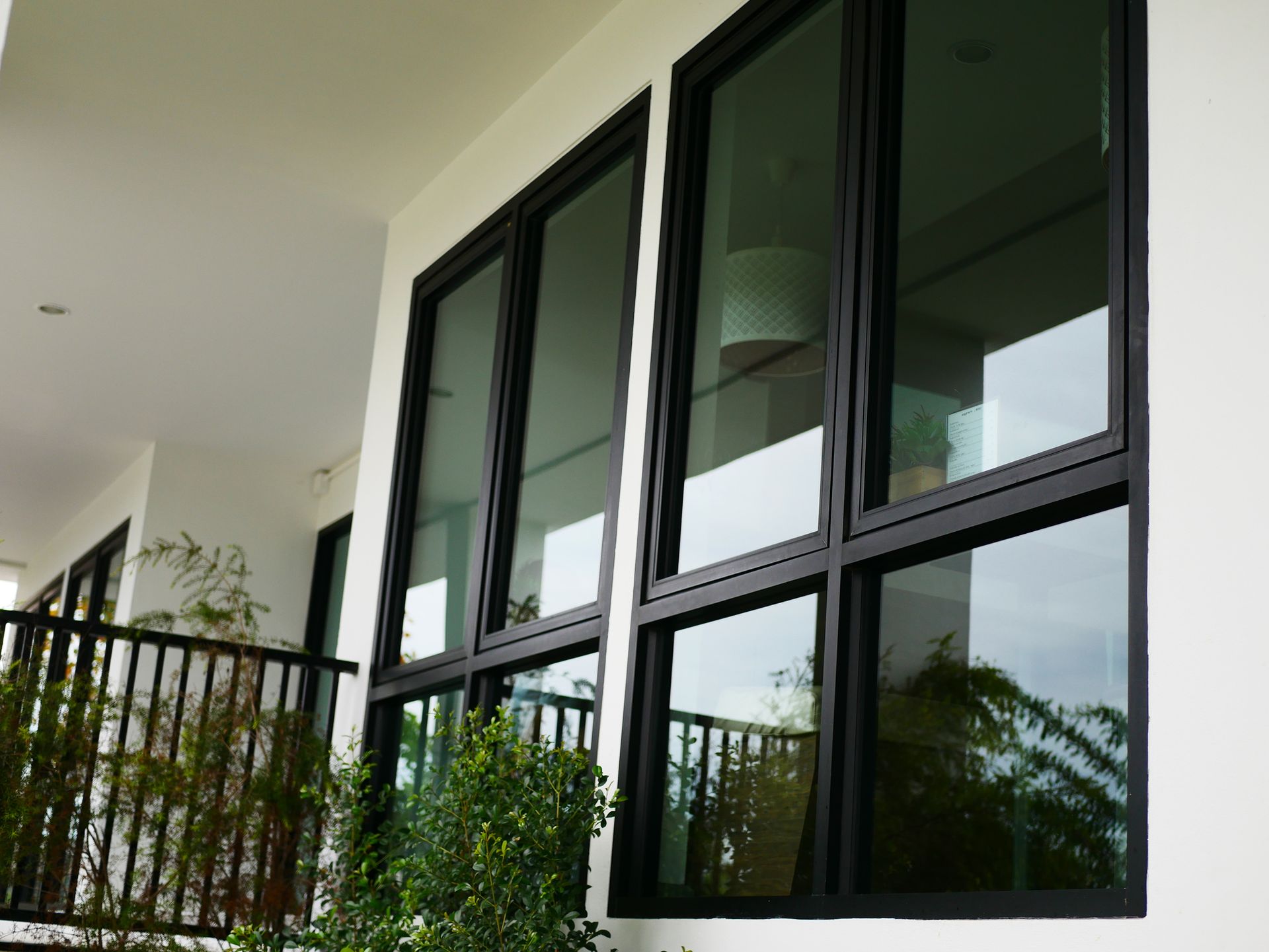 Black-framed windows on a white building, reflecting the outside. A small balcony and plants are visible.
