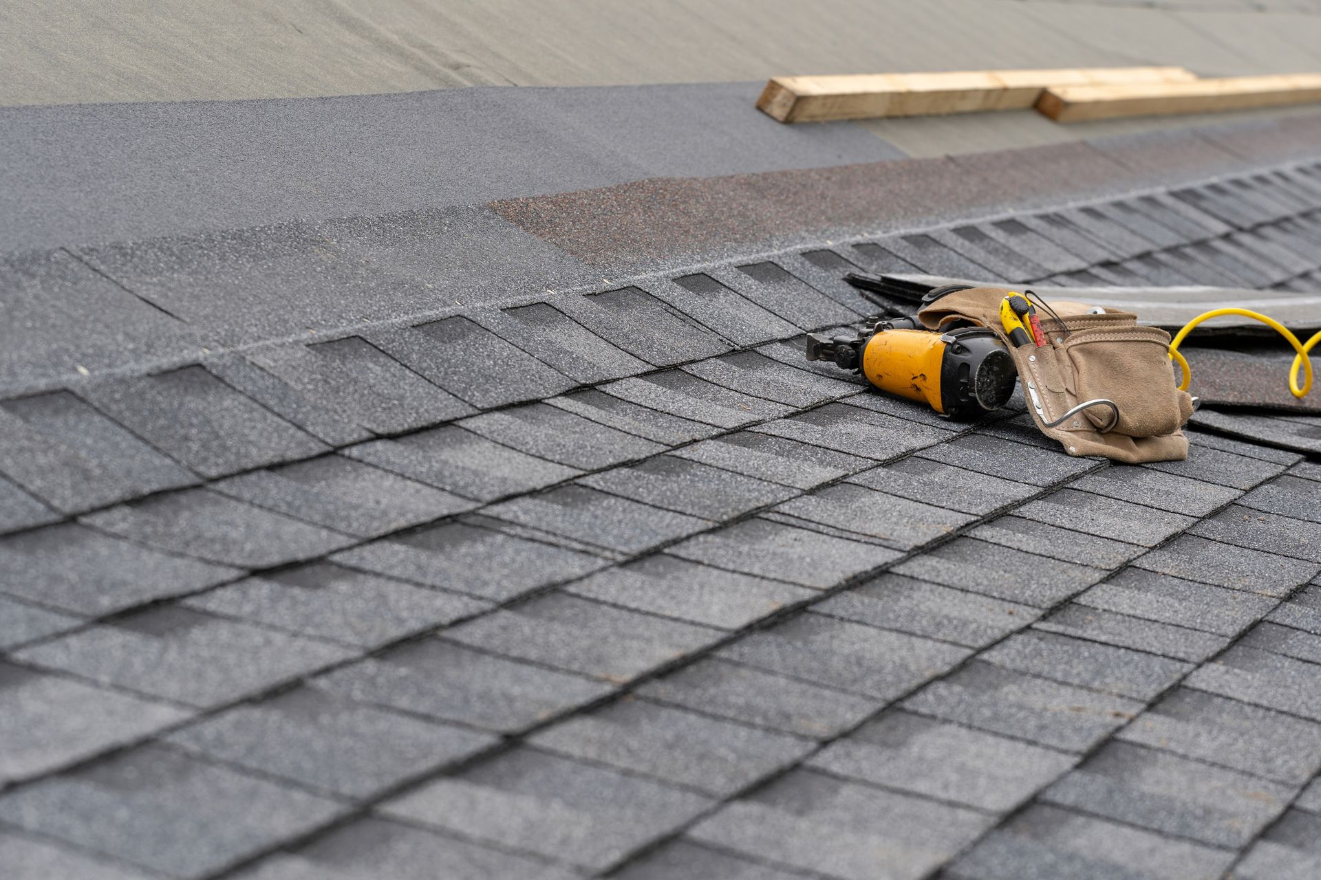 Roofing work in progress: a nail gun and tool belt sit atop gray asphalt shingles.