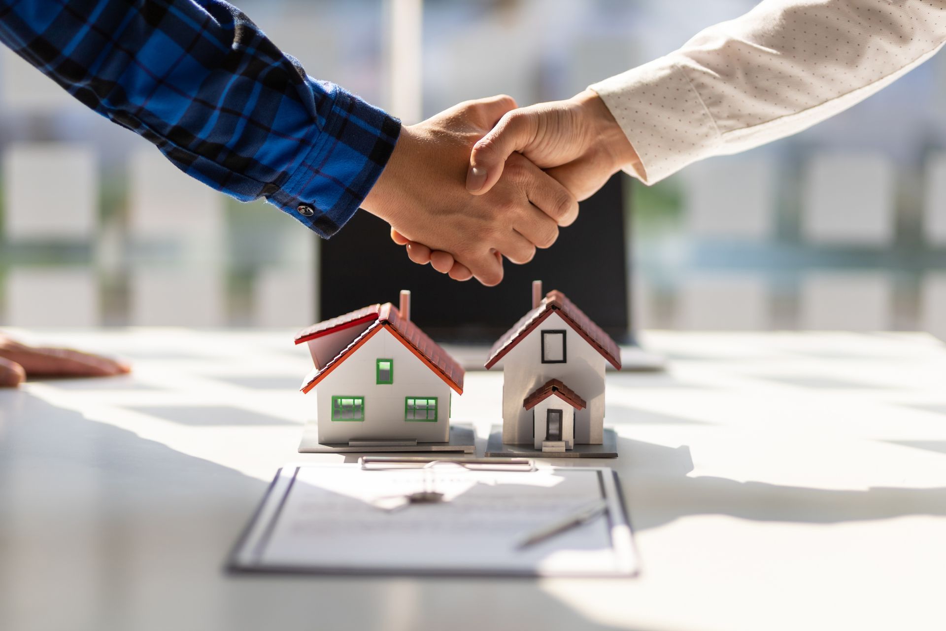 Two people shaking hands over a table with miniature houses and a real estate contract.