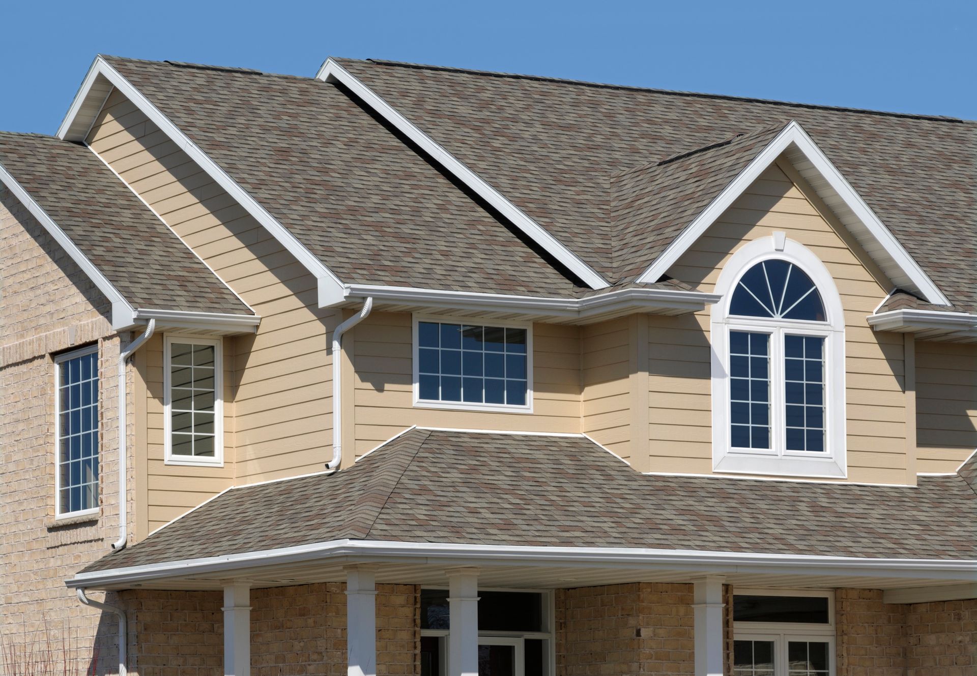 Tan-colored house with a multi-angled roof. Windows with white frames are visible, along with beige siding.