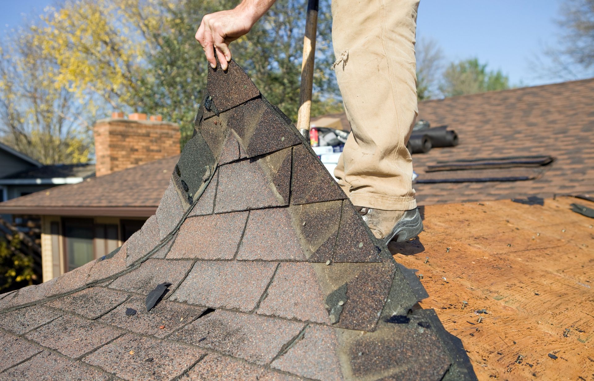 A roofer removes a worn shingle from a chimney's cap on a brown shingled roof.