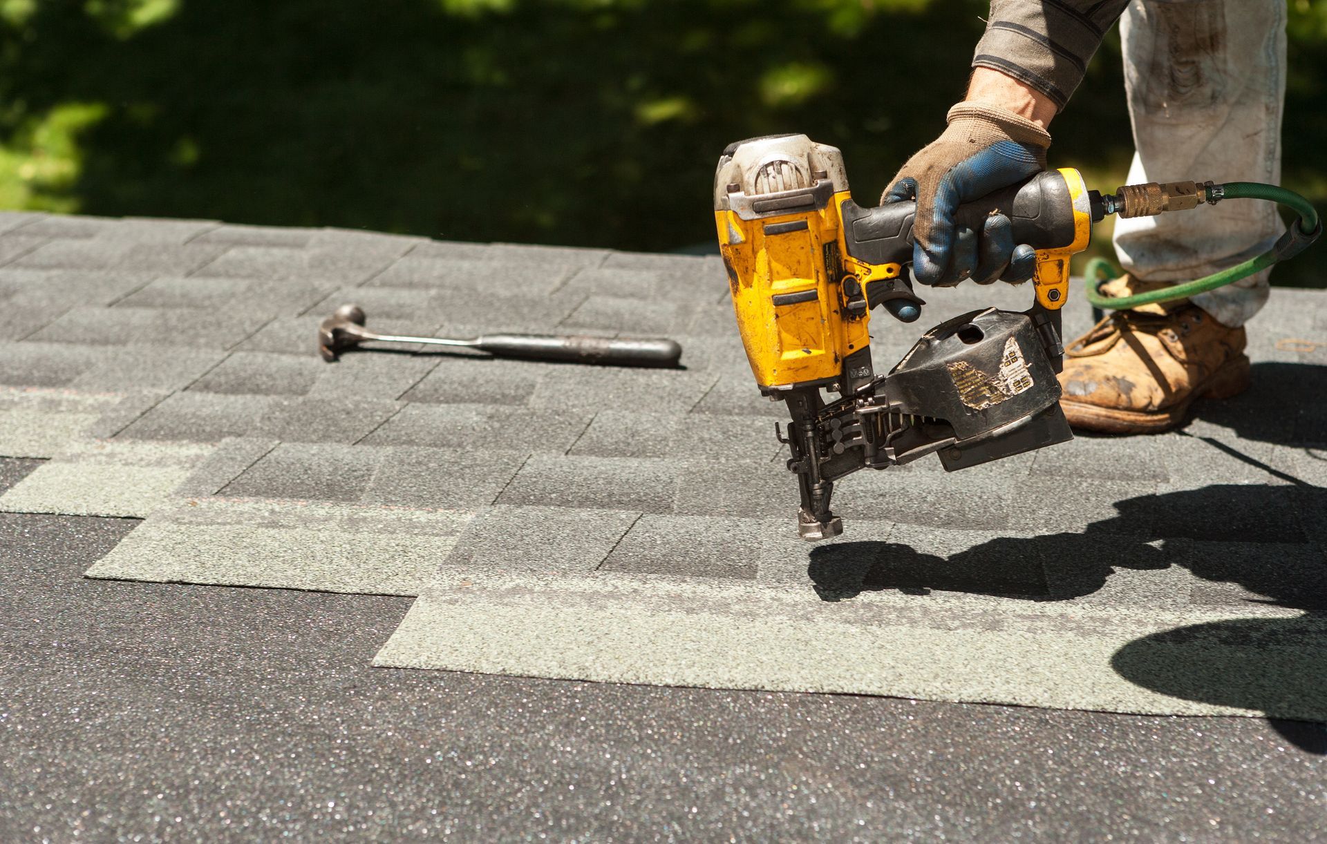 A worker uses a nail gun to install asphalt shingles on a roof. A hammer sits nearby.