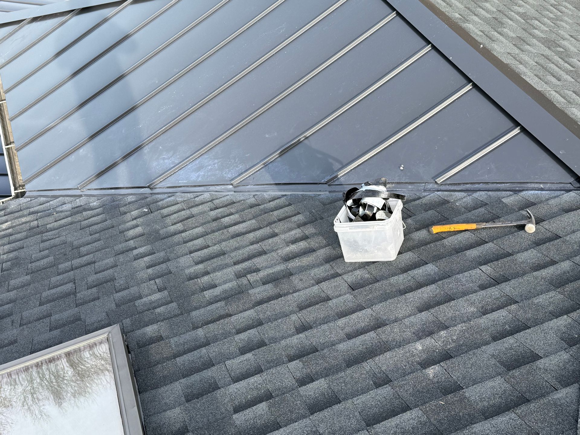 View from above, a roof with dark gray shingles and a gray metal section. Tools sit on the roof.