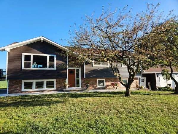 Brown and gray two-story house with a green lawn, clear blue sky, and a tree in front.