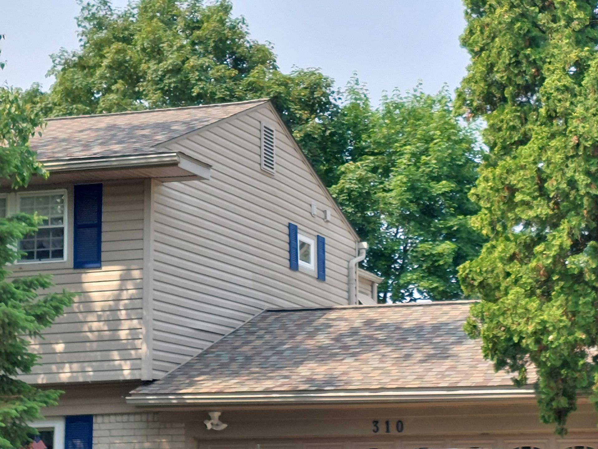 Tan house with blue shutters, surrounded by green trees.